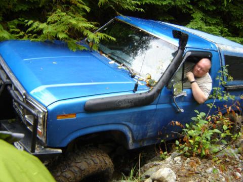1985 Ford Bronco - A blue Ford Bronco is parked off-road with a snorkel intake visible on the driver's side. The driver is leaning out the window looking down.