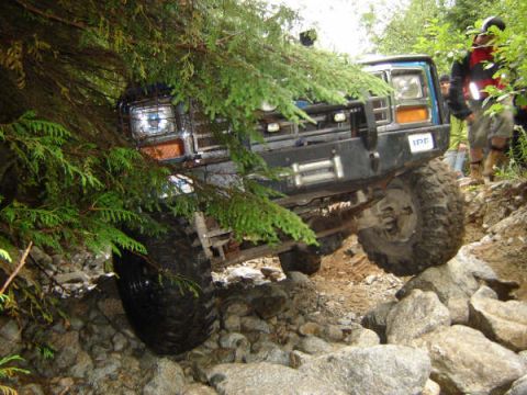 1985 Ford Bronco - A blue off-road SUV navigates a rocky, wooded trail, with its front tire climbing over large boulders. The vehicle features a winch and aft…