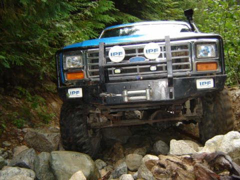 1985 Ford Bronco - A blue Ford Bronco is navigating a rocky, wooded trail. The front of the vehicle features a black bull bar with IPF lights and a winch.