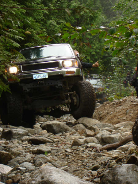 1985 Ford Bronco - A black off-road pickup truck with large tires navigates a rocky, wooded trail. The truck's headlights are on, and a person is visible in t…