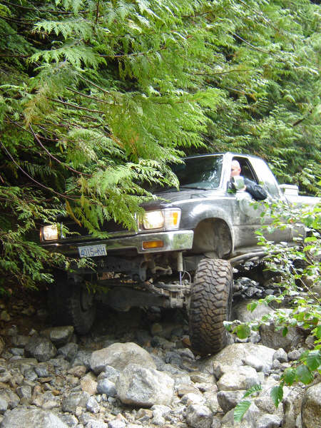 1985 Ford Bronco - A silver 4x4 truck navigates a rocky, wooded trail, with its front tire angled upwards over a large boulder. The license plate reads "4015-…