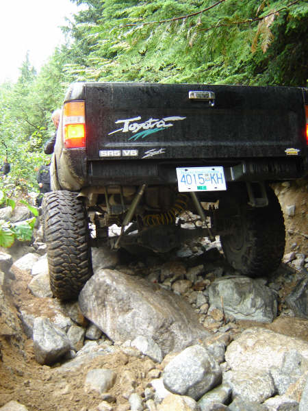 1985 Ford Bronco - The rear of a black Toyota pickup truck is shown navigating a rocky trail. The truck has a license plate reading "4015 KH" and large off-ro…