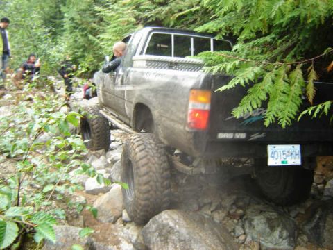 1985 Ford Bronco - A dark-colored pickup truck with large off-road tires navigates a rocky, wooded trail. The vehicle's rear license plate reads "4015 KH".