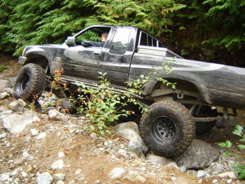 1985 Ford Bronco - A black pickup truck is navigating a rocky, off-road trail surrounded by dense green foliage. The truck's large tires are positioned over r…