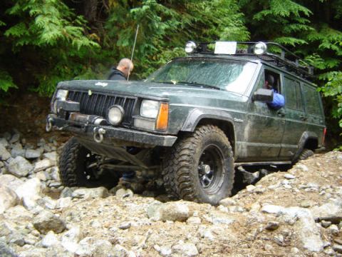 1985 Ford Bronco - A dark green Jeep Cherokee XJ is navigating a rocky, off-road trail with dense green foliage in the background. The vehicle is equipped wit…