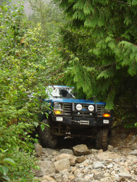 1985 Ford Bronco - A blue 1985 Ford Bronco is navigating a rocky, wooded trail. The front of the vehicle is visible, showing off-road lights and a winch.