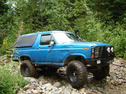 1985 Ford Bronco - A blue Ford Bronco is shown from a 3/4 front angle driving over a rocky, wooded terrain. The vehicle has large off-road tires, a snorkel, a…