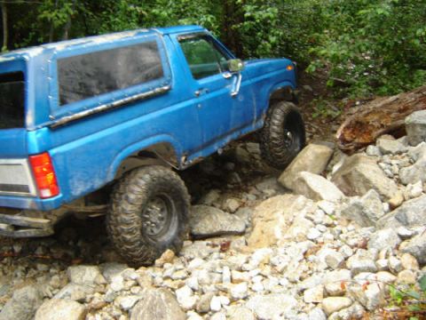 1985 Ford Bronco - A blue Ford Bronco is navigating a rocky, off-road trail with large tires and a lifted suspension. The vehicle is angled to the right, show…