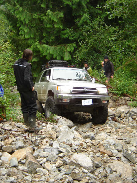 1985 Ford Bronco - A silver SUV navigates a rocky, wooded trail with several people standing nearby.