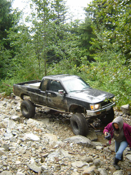 1985 Ford Bronco - A black Toyota pickup truck is navigating a rocky, off-road trail surrounded by dense green foliage. A person in a hooded jacket is walking…
