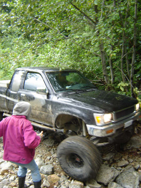 1985 Ford Bronco - A black Toyota pickup truck is navigating a rocky, wooded trail. A person in a pink jacket and grey hoodie is standing near the front of th…