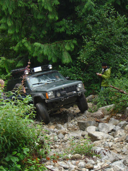 1985 Ford Bronco - A dark-colored Jeep Cherokee is navigating a rocky, off-road trail surrounded by dense green foliage. A person in a green jacket stands to…