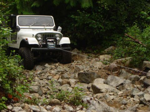 1985 Ford Bronco - A white Jeep CJ is parked on a rocky, wooded trail, with a winch mounted on the front bumper. A tow rope extends from the winch across the…