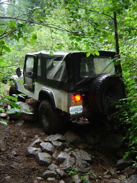 1985 Ford Bronco - A white and black Jeep Wrangler with a soft top is navigating a rocky, muddy trail in a dense forest.