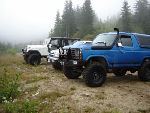 1985 Ford Bronco - A blue Ford Bronco with a snorkel intake is parked off-road on a dirt path with a white Jeep and a black SUV behind it. The vehicles are su…