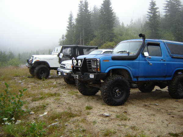 Blue 1985 Ford Bronco with snorkel, parked off-road with other vehicles.