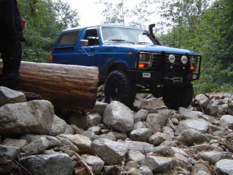 1985 Ford Bronco - A blue 1980s Ford Bronco is navigating a rocky, wooded trail, with a large log in the foreground.
