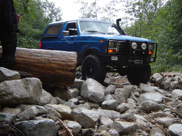 Blue 1980s Ford Bronco off-roading over rocks and a log.