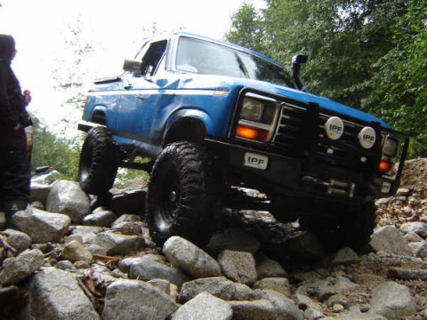 1985 Ford Bronco - A lifted blue Ford Bronco is navigating a rocky, off-road trail with a person standing to the left. The vehicle features large off-road tir…