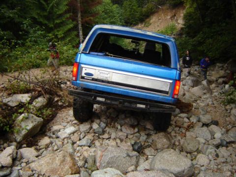 1985 Ford Bronco - A blue Ford Bronco is navigating a rocky, wooded trail, with two people standing on the side of the trail behind it.
