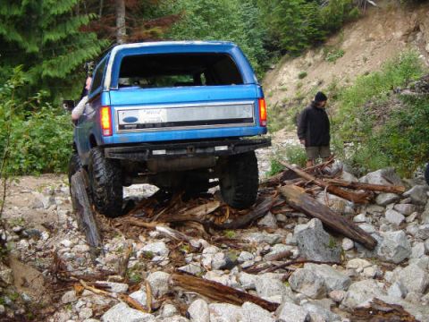 1985 Ford Bronco - A blue Ford Bronco is navigating a rocky, wooded trail, with its rear tire resting on a large log. A person in a dark jacket and shorts sta…