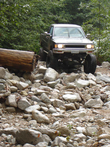 1985 Ford Bronco - A lifted, silver pickup truck navigates a rocky, off-road trail with a large log to its left. The truck is angled towards the viewer, showi…