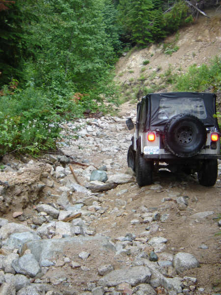 1985 Ford Bronco - A white Jeep Wrangler with a soft top navigates a rocky, off-road trail in a wooded area. The vehicle is seen from the rear-left, ascending…