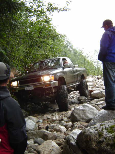 1985 Ford Bronco - A silver pickup truck navigates a rocky, off-road trail with a person standing on the rocks to the right, observing.