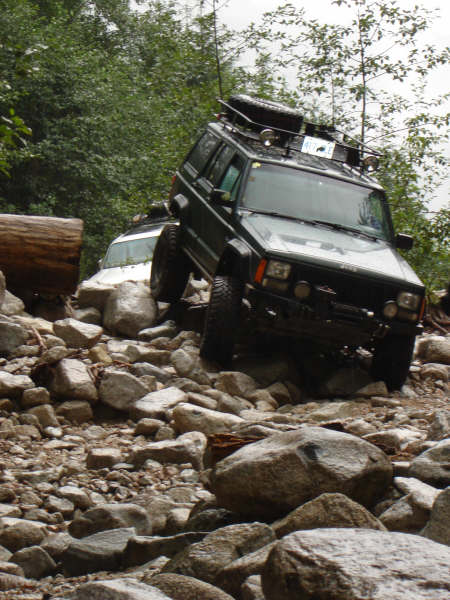 1985 Ford Bronco - A dark colored Jeep Cherokee is navigating a rocky, off-road trail. Another white SUV is partially visible behind it.