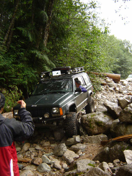 1985 Ford Bronco - A dark green Jeep Cherokee is navigating a rocky, wooded trail. The vehicle is equipped with a roof rack and large off-road tires.