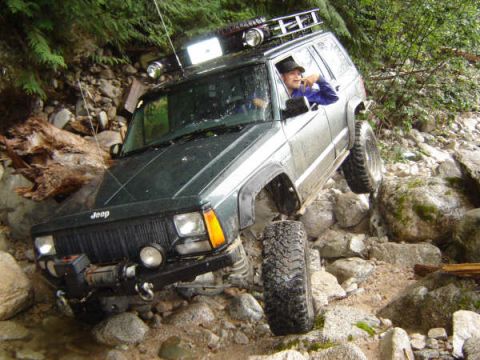 1985 Ford Bronco - A dark green Jeep Cherokee is navigating a rocky, off-road trail. The driver is visible through the window, wearing a hat and looking towar…