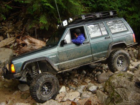 1985 Ford Bronco - A dark green Jeep Cherokee is navigating a rocky trail with large boulders and trees surrounding it. The vehicle has a roof rack and large…