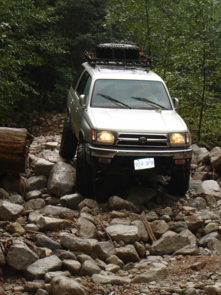 1985 Ford Bronco - A silver SUV is navigating a rocky, off-road trail surrounded by dense trees. The vehicle has a roof rack with a spare tire and its headlig…