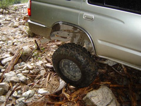 1985 Ford Bronco - The rear passenger side of a silver SUV is shown at an angle, with its tire resting on rocks and debris on a rough, unpaved surface.