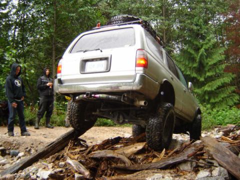 1985 Ford Bronco - A silver SUV is navigating a rocky, wooded trail, with its front tire elevated on a log. Two people in hooded sweatshirts stand behind the…