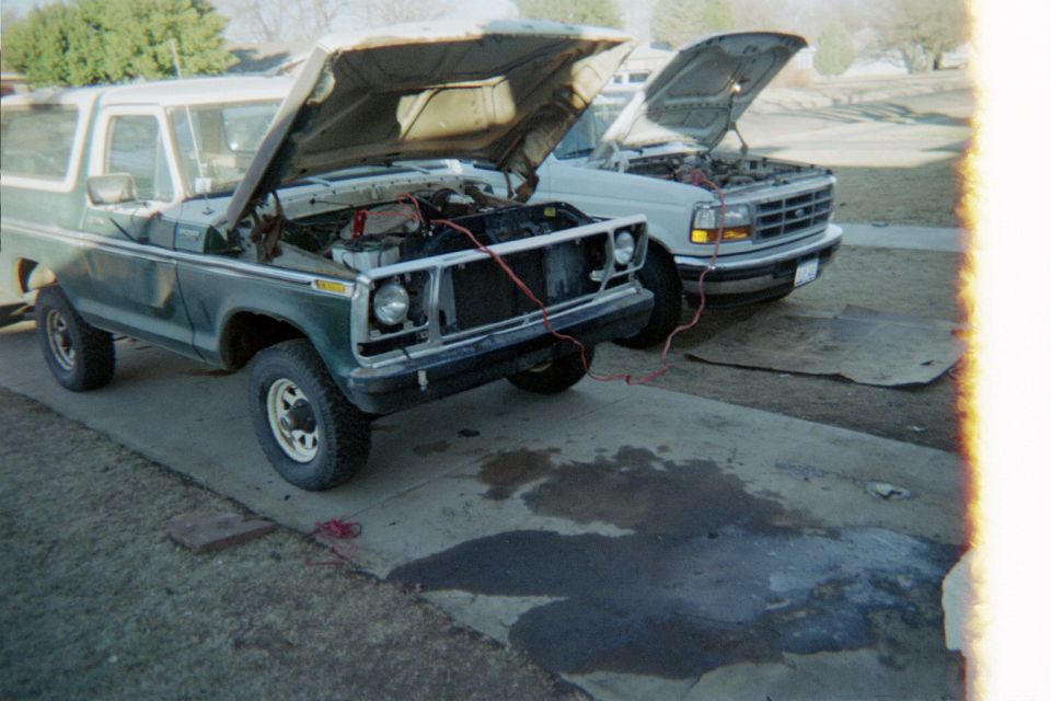 Green 1978 Ford Bronco with hood open, being jump-started by a white Ford F-Series truck
