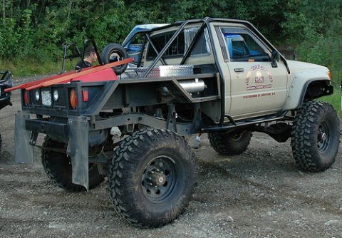 1985 Ford Bronco - A heavily modified, lifted pickup truck with large off-road tires and a custom flatbed is parked on a gravel surface. The truck has a roll…