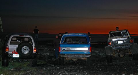 1985 Ford Bronco - Three off-road vehicles are parked on a rocky outcrop at dusk. The vehicle on the right is a black pickup truck with a flatbed and a roll c…