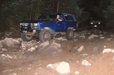 1985 Ford Bronco - A blue Ford Bronco is being towed by another vehicle on a rocky, wooded trail at night. The Bronco has a brush guard and large off-road tir…