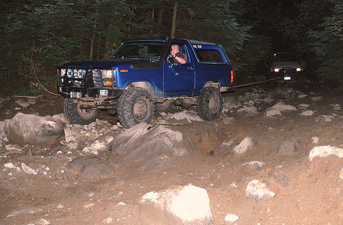 Blue Ford Bronco on a rocky trail at night, being towed by another vehicle.