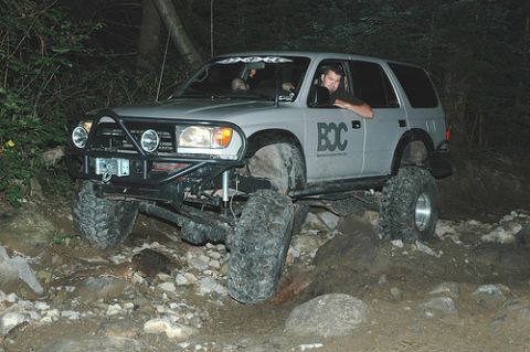 1985 Ford Bronco - A lifted, silver SUV with large off-road tires navigates a rocky trail at night. The driver is visible behind the wheel, and the vehicle ha…