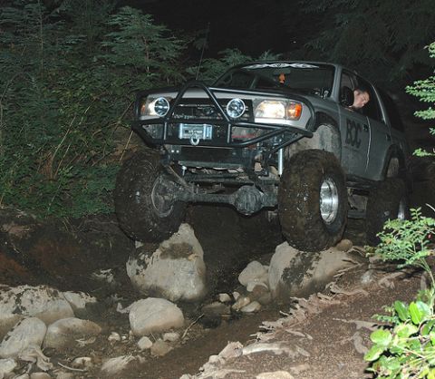 1985 Ford Bronco - A lifted, silver SUV with large off-road tires navigates a rocky trail at night. The vehicle has a custom front bumper with a winch and aux…