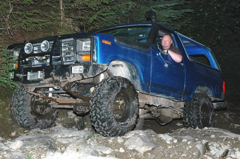1985 Ford Bronco - A blue 1985 Ford Bronco is navigating a rocky trail with large off-road tires. A man is visible driving the vehicle, which has a snorkel an…