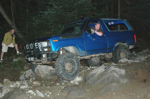 1985 Ford Bronco - A blue 1980s Ford Bronco is navigating a rocky, wooded trail at night. A man is driving, and another man stands beside the vehicle.