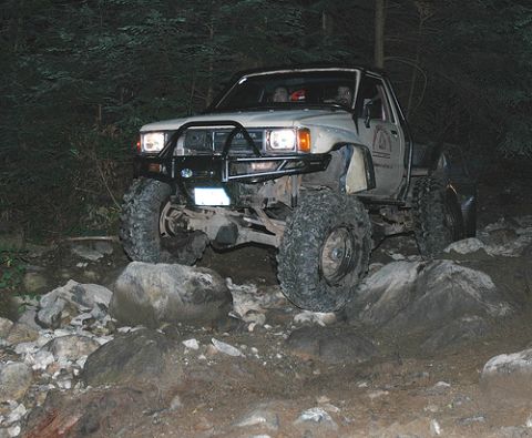 1985 Ford Bronco - A lifted, light-colored pickup truck with large off-road tires navigates a rocky, wooded trail at night. The truck has a black brush guard…