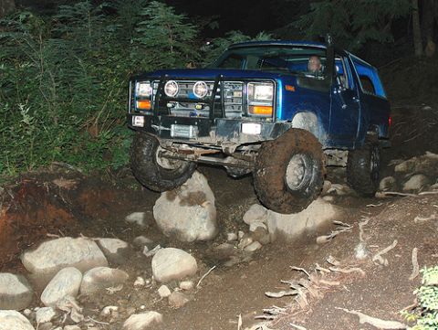 1985 Ford Bronco - A blue 1985 Ford Bronco is navigating a rocky, wooded trail at night. The vehicle is angled with its front right tire on a large rock, show…