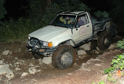 1985 Ford Bronco - A lifted, white pickup truck with large off-road tires navigates a rocky, wooded trail at night. The truck has a custom front bumper and ro…