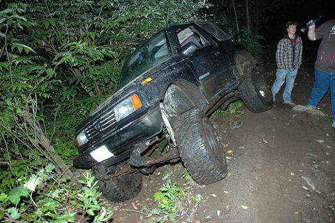 1985 Ford Bronco - A black Suzuki Samurai is tilted on its side on a muddy trail, with its front passenger side wheel elevated and its rear driver's side whee…