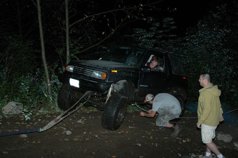 1985 Ford Bronco - A black SUV is stuck in the mud at night, with two people working to free it using a tow strap attached to the front bumper. Another person…