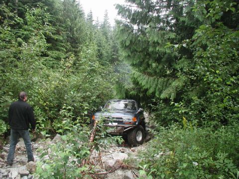 1985 Ford Bronco - A dark-colored SUV with a custom front bumper navigates a rocky, wooded trail. A person in a black jacket stands on the left side of the tr…